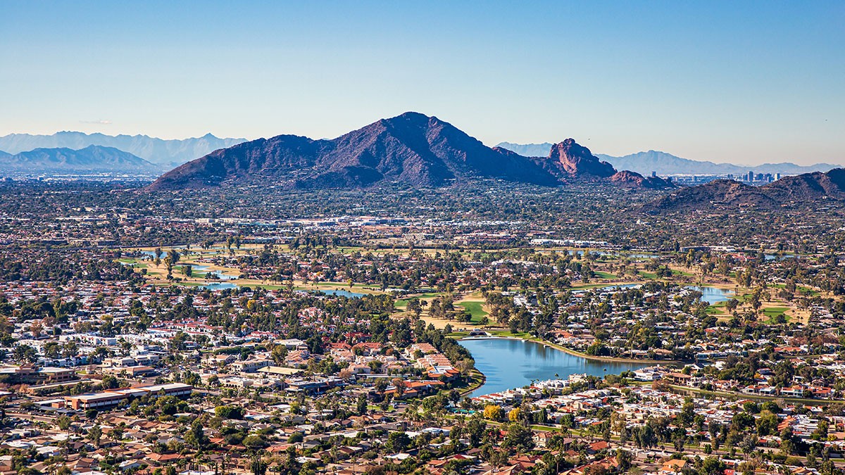 Aerial view of a city with scattered buildings, green spaces, and a central lake, set against a backdrop of mountains under a clear blue sky.