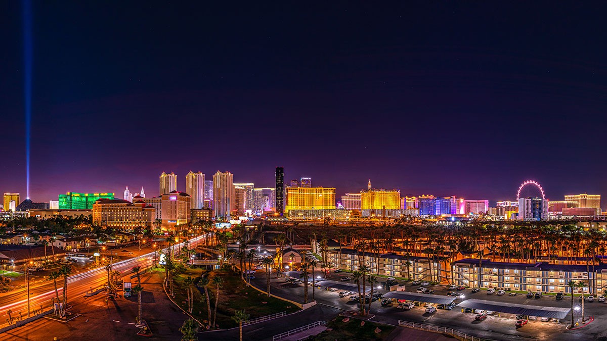 Night view of the Las Vegas Strip with illuminated hotels, casinos, a Ferris wheel, bright lights, and car trails on the road.