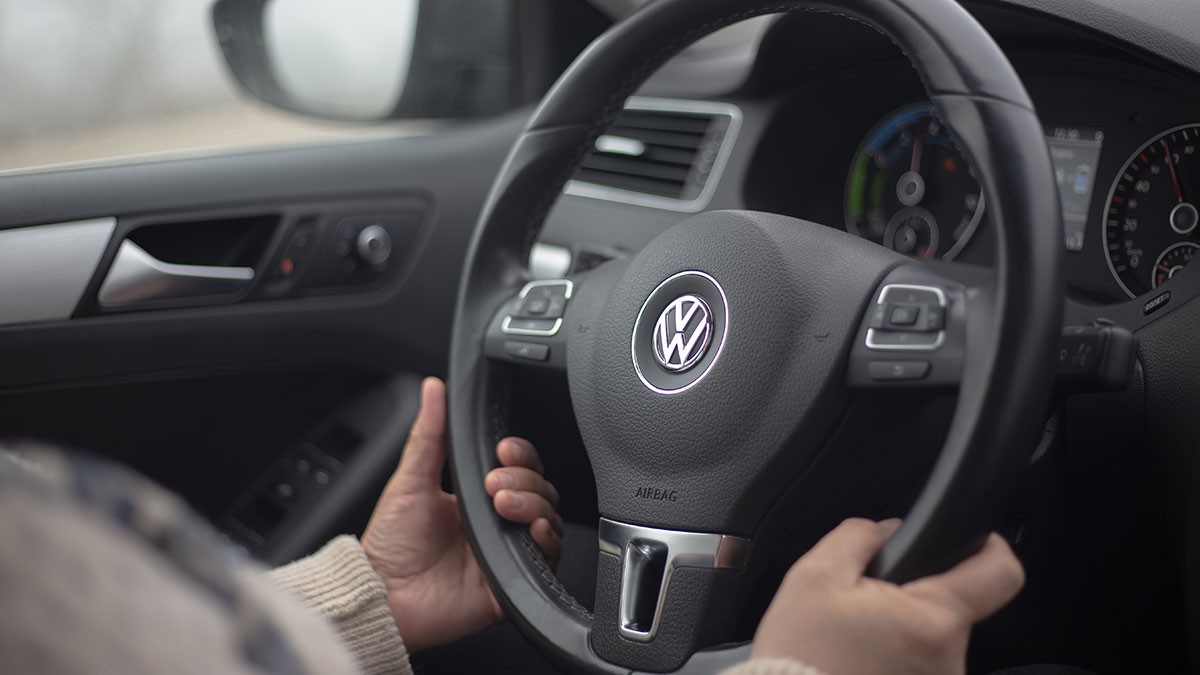 Person holding a Volkswagen steering wheel inside a car, with dashboard and controls visible in the background.