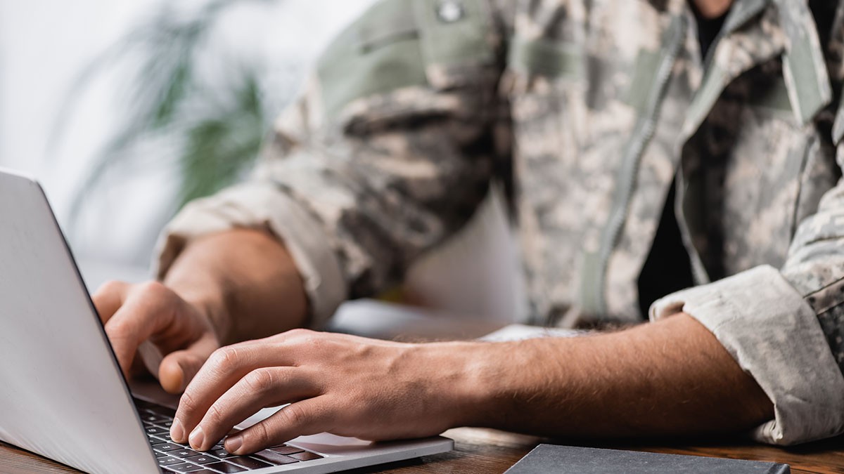 Person in camouflage using a laptop at a desk, with plants in the background.
