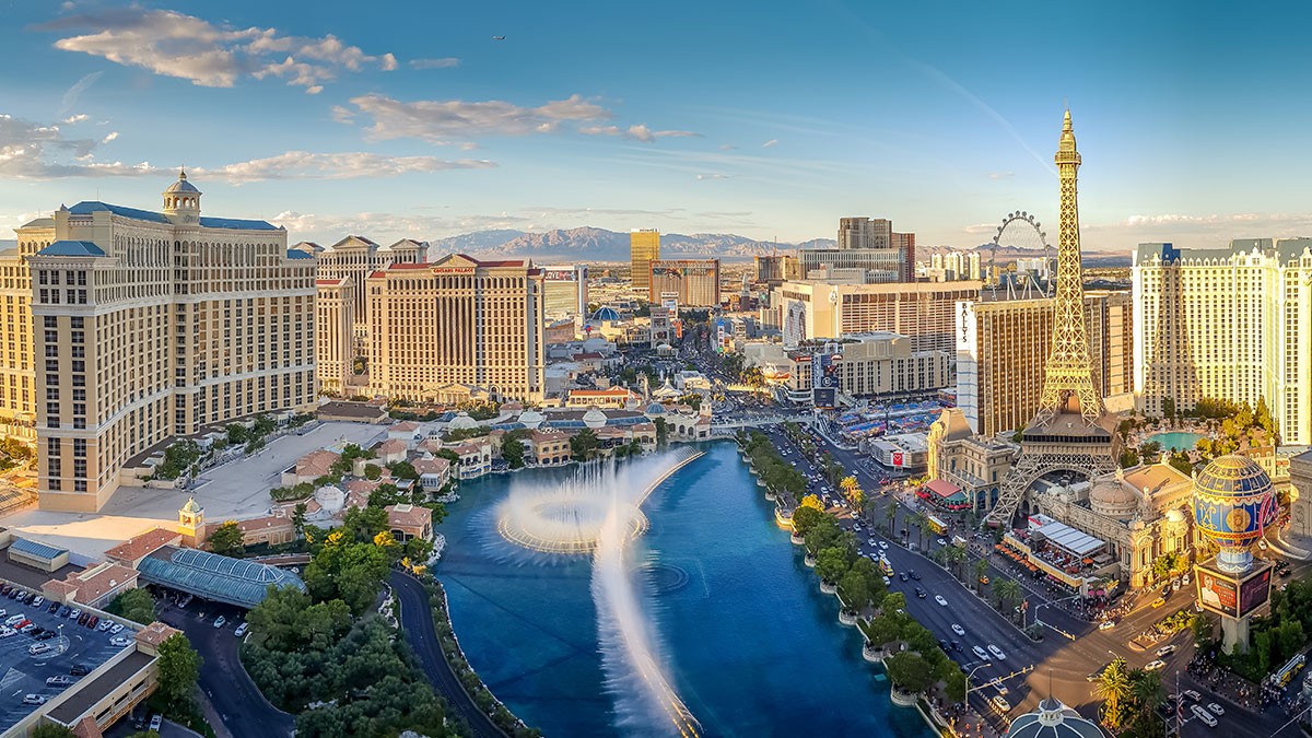 Aerial view of Las Vegas Strip with fountains, hotels, and replica Eiffel Tower under a clear blue sky.