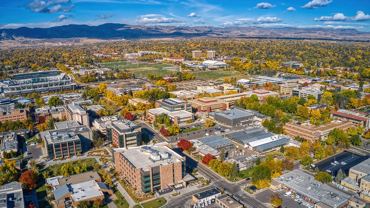 Aerial view of a sprawling university campus surrounded by autumn trees, with mountains in the background under a blue sky with clouds.