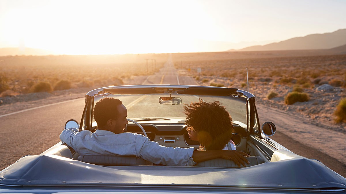 Two people in a convertible on an open desert road, enjoying a scenic sunset.