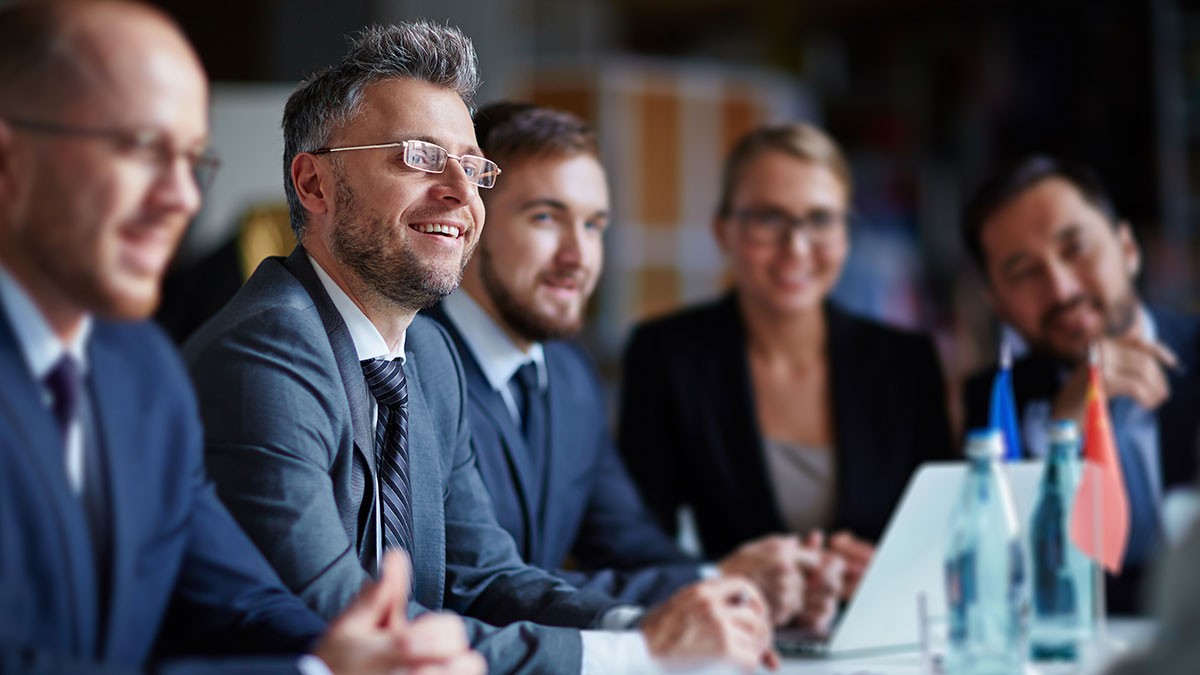 Group of five business professionals in suits sitting at a conference table, engaged in discussion, with laptops and flags visible.