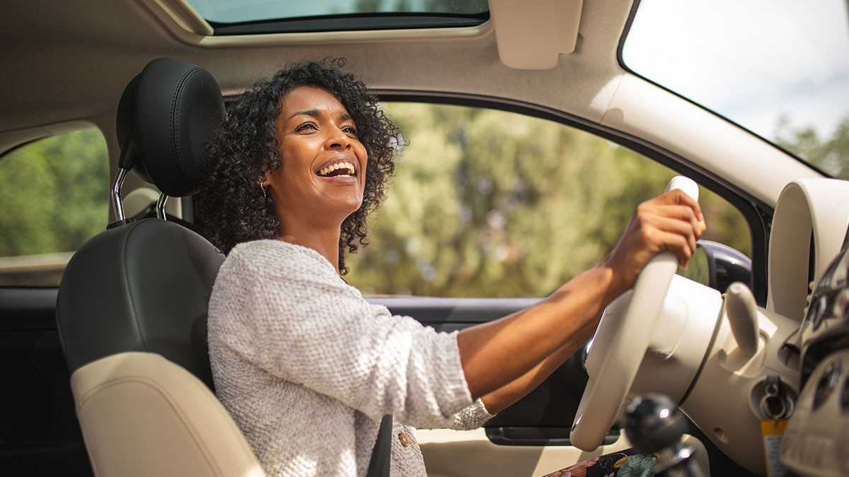 Smiling woman with curly hair driving a car, sunroof open, and trees visible outside.