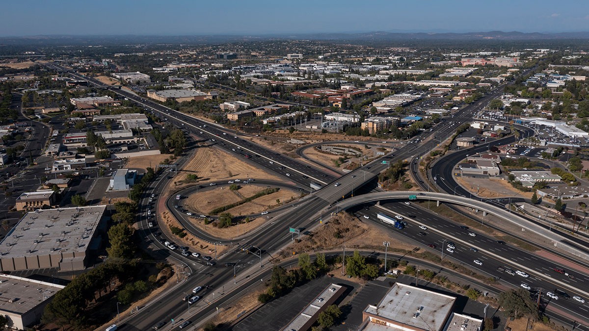 Aerial view of a sprawling highway interchange with looping ramps, multiple lanes of traffic and surrounding commercial buildings and dry landscape.