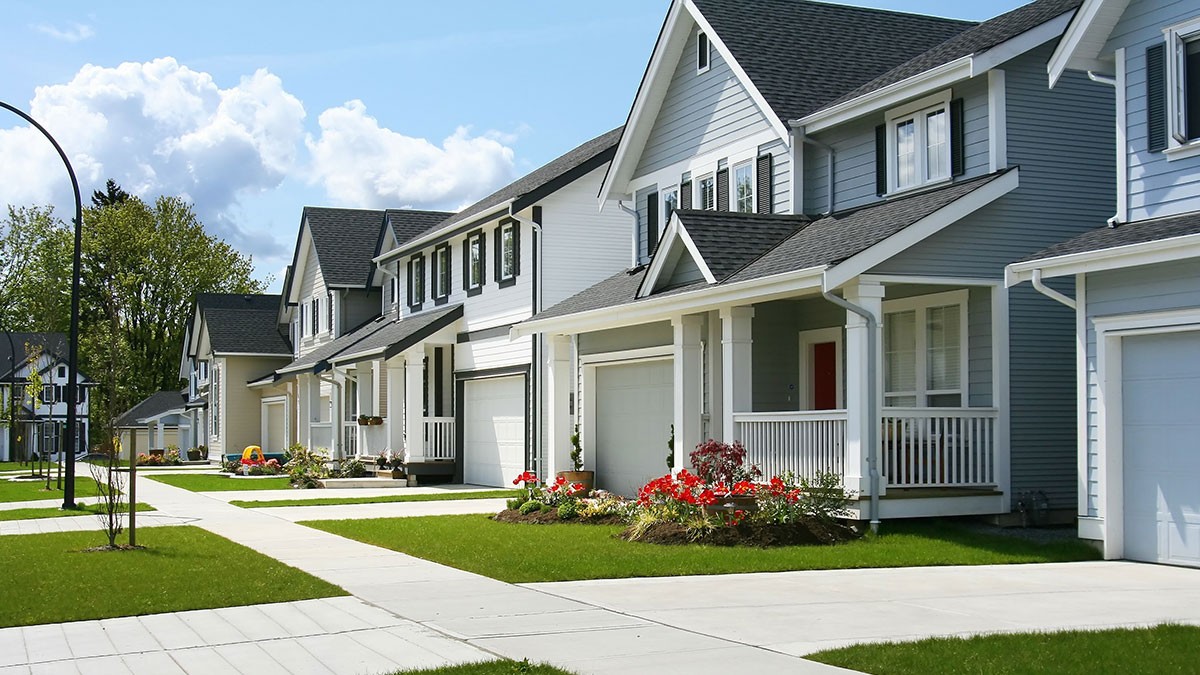 A row of modern suburban houses with manicured lawns, white facades, and grey roofs, under a partly cloudy sky.