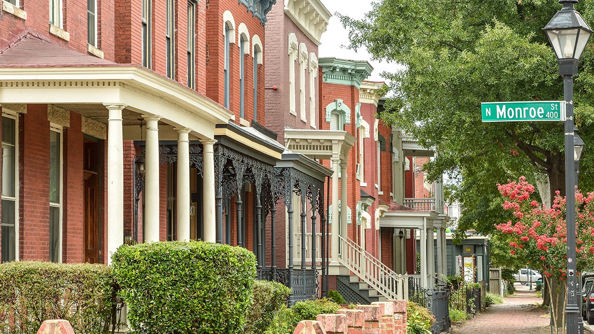 A row of historic brick townhouses with ornate porches line a leafy street, featuring a green sign reading "N Monroe St."