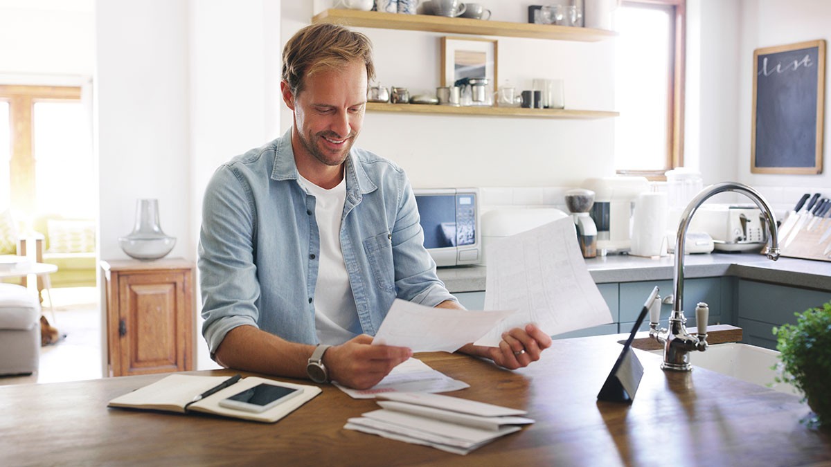 A smiling man sits at a kitchen counter, holding paperwork. A tablet, notebook, and phone are on the counter. The kitchen is modern and bright.
