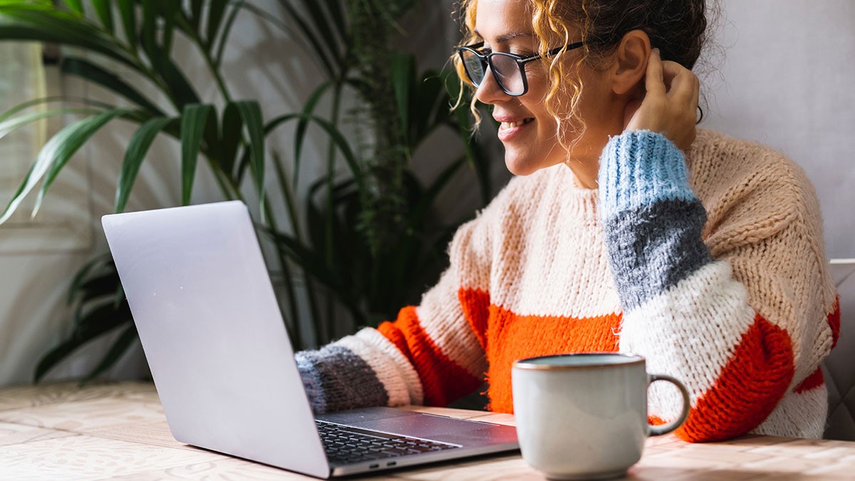 A woman in glasses and a colorful sweater uses a laptop at a table with a mug, next to a large plant.