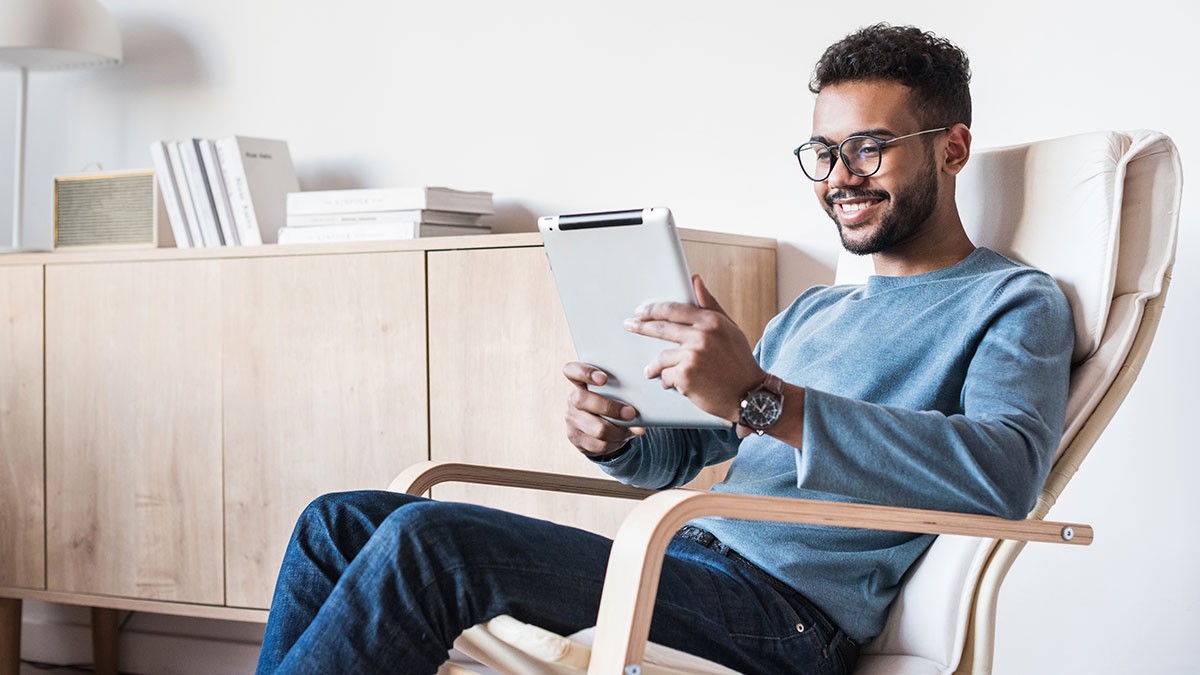 Man sitting in a chair, smiling while using a tablet. He wears glasses and a blue sweater, with a wooden cabinet and books in the background.