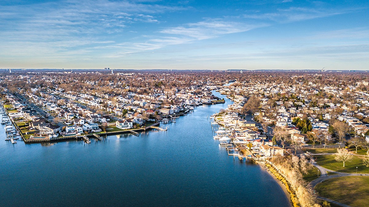 Aerial view of a coastal town with numerous houses lining the shores of a winding waterway, under a partly cloudy blue sky.