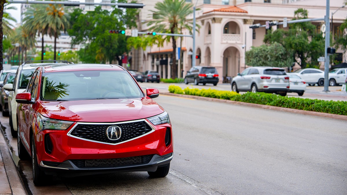 A red SUV parked on a city street lined with palm trees and buildings, with other cars visible in the background.
