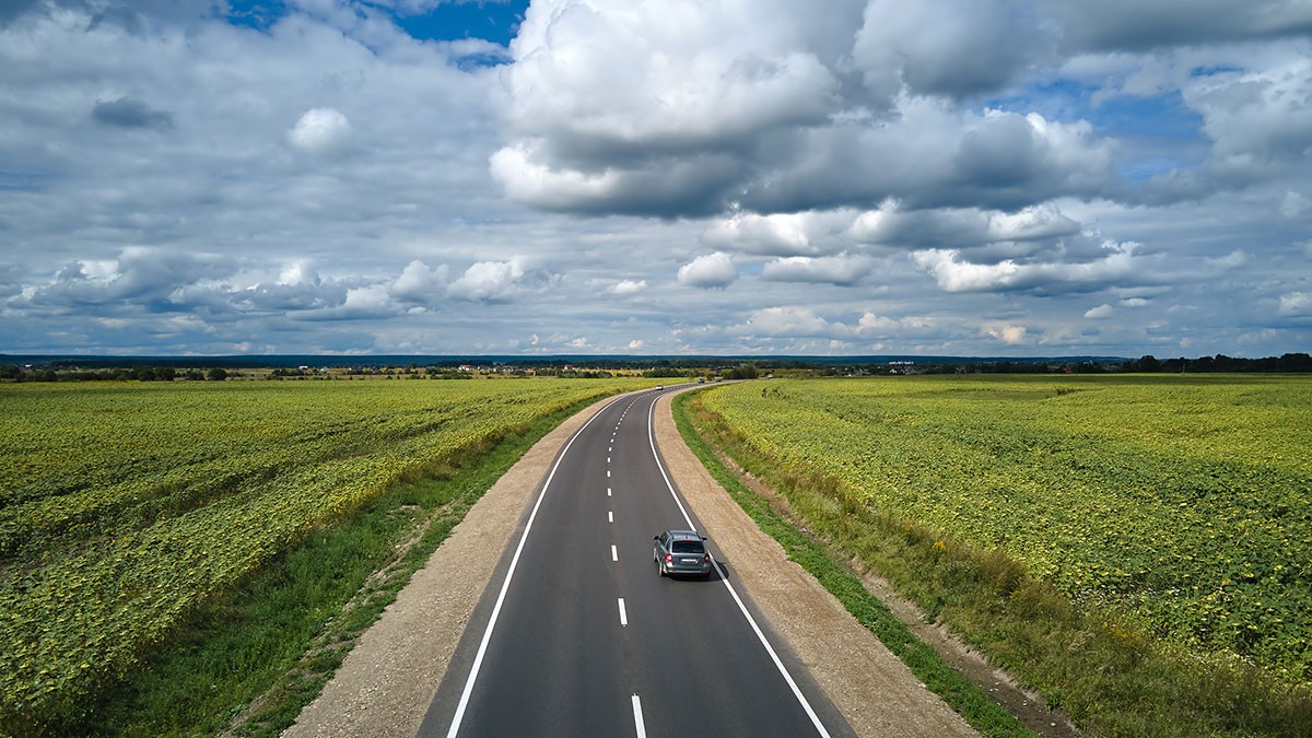 A car drives down a long, empty road flanked by vast green fields under a cloudy sky.