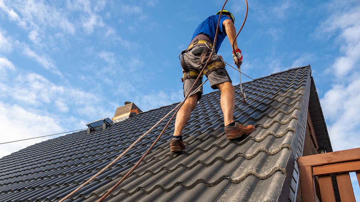 A person wearing safety gear and shorts is working on a sloped roof with dark tiles, set against a partly cloudy sky.