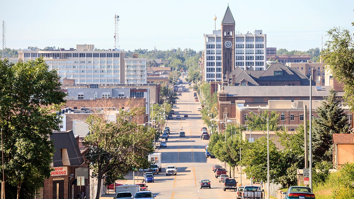 A city street with cars, lined by trees and buildings, leading to a distant clock tower under a clear blue sky.