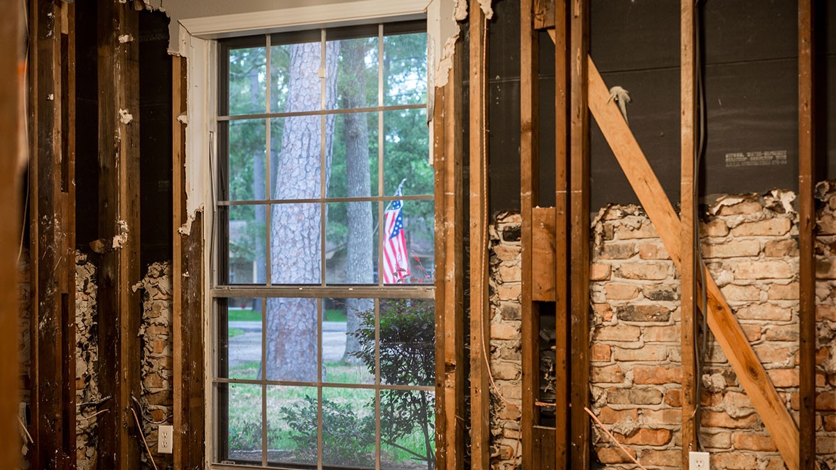 Partially stripped interior wall revealing studs and brick, with a window showing a tree and an American flag outside.