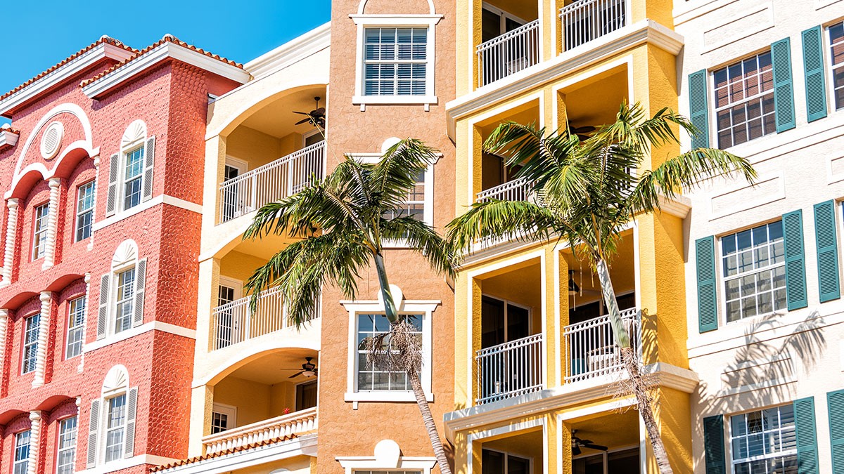 Colorful Mediterranean-style apartment buildings with balconies and palm trees in front, under a clear blue sky.