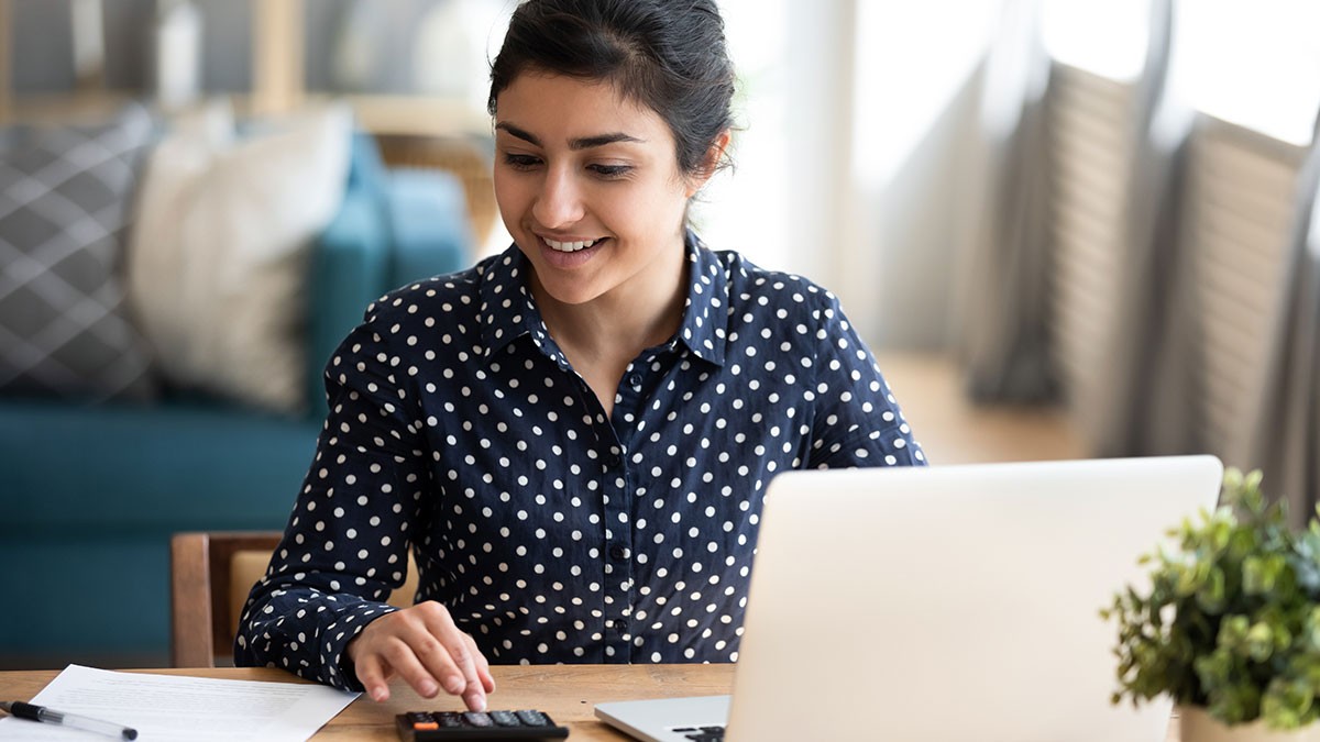 A woman in a polka dot shirt uses a calculator while working on a laptop at a desk, with papers nearby and a plant in the foreground.