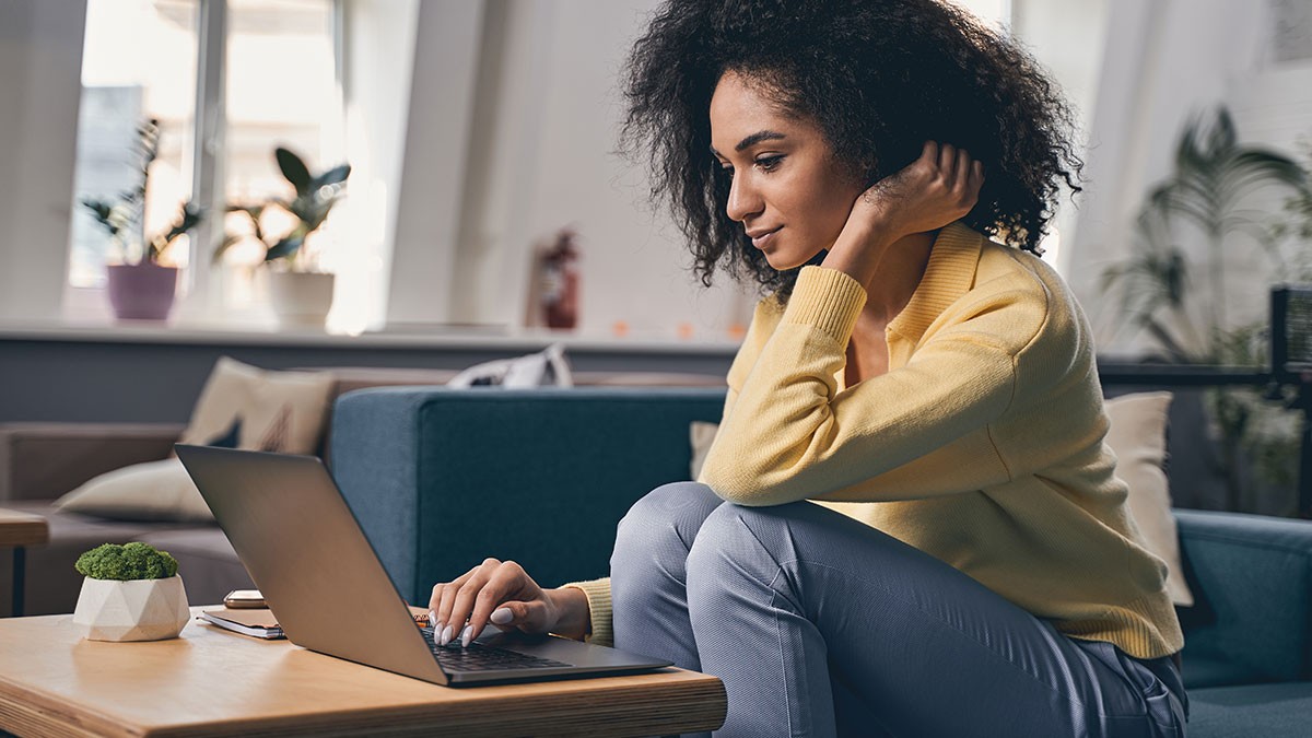 Woman in a yellow sweater working on a laptop, seated comfortably in a cozy room with soft lighting and modern decor.