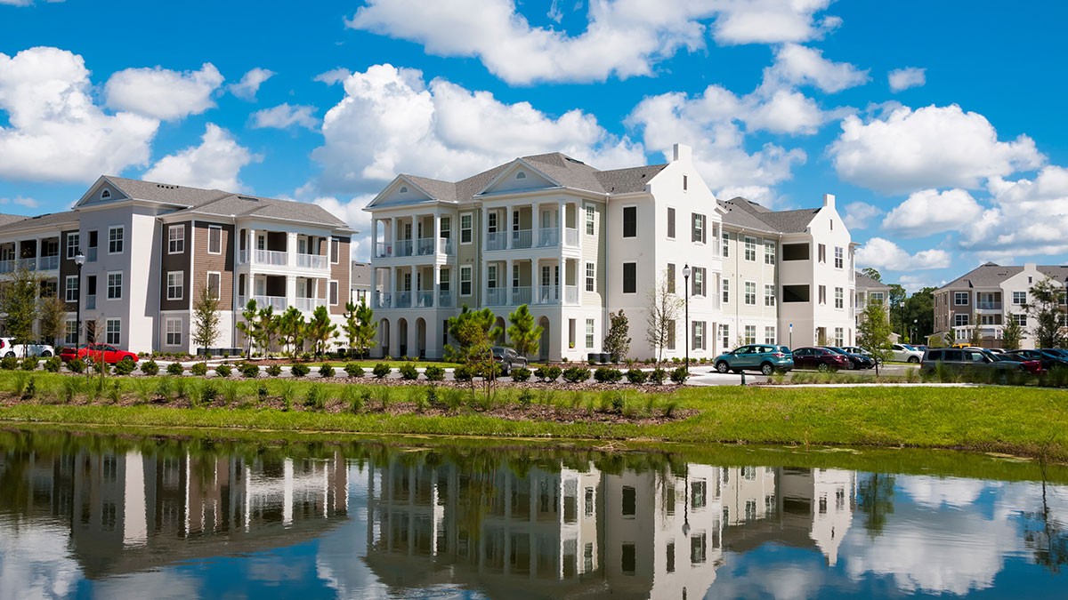 Apartment buildings with balconies by a reflective pond under a partly cloudy blue sky.