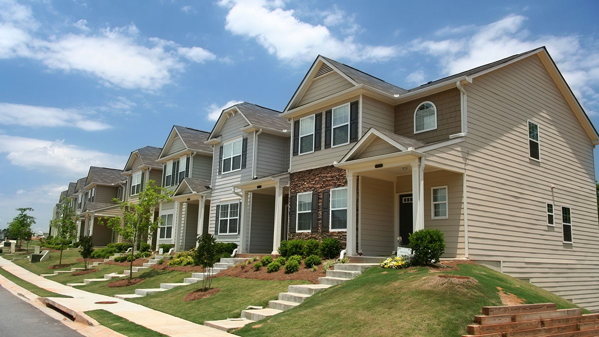 A row of modern suburban houses with sloped lawns, light siding, and small porches on a sunny day.