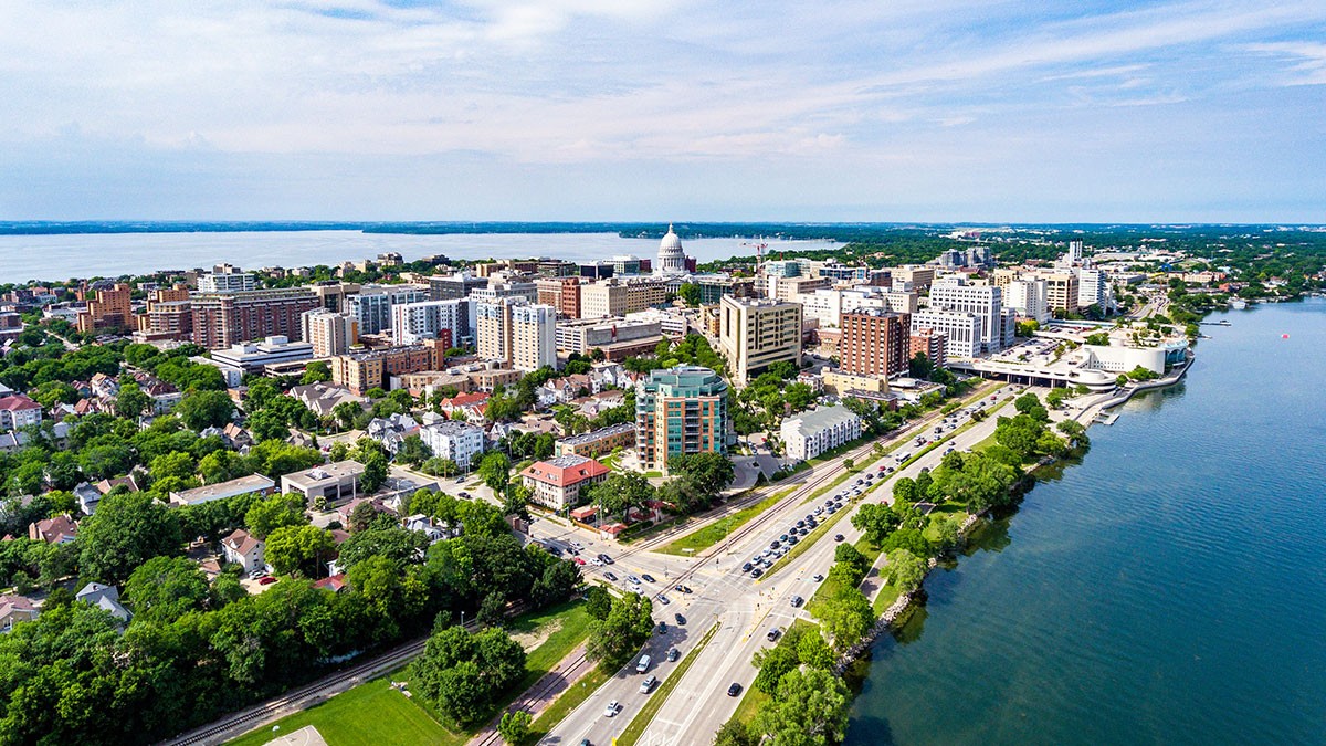 Aerial view of a cityscape with a mix of buildings, roads, and greenery, bordered by a large body of water under a partly cloudy sky.