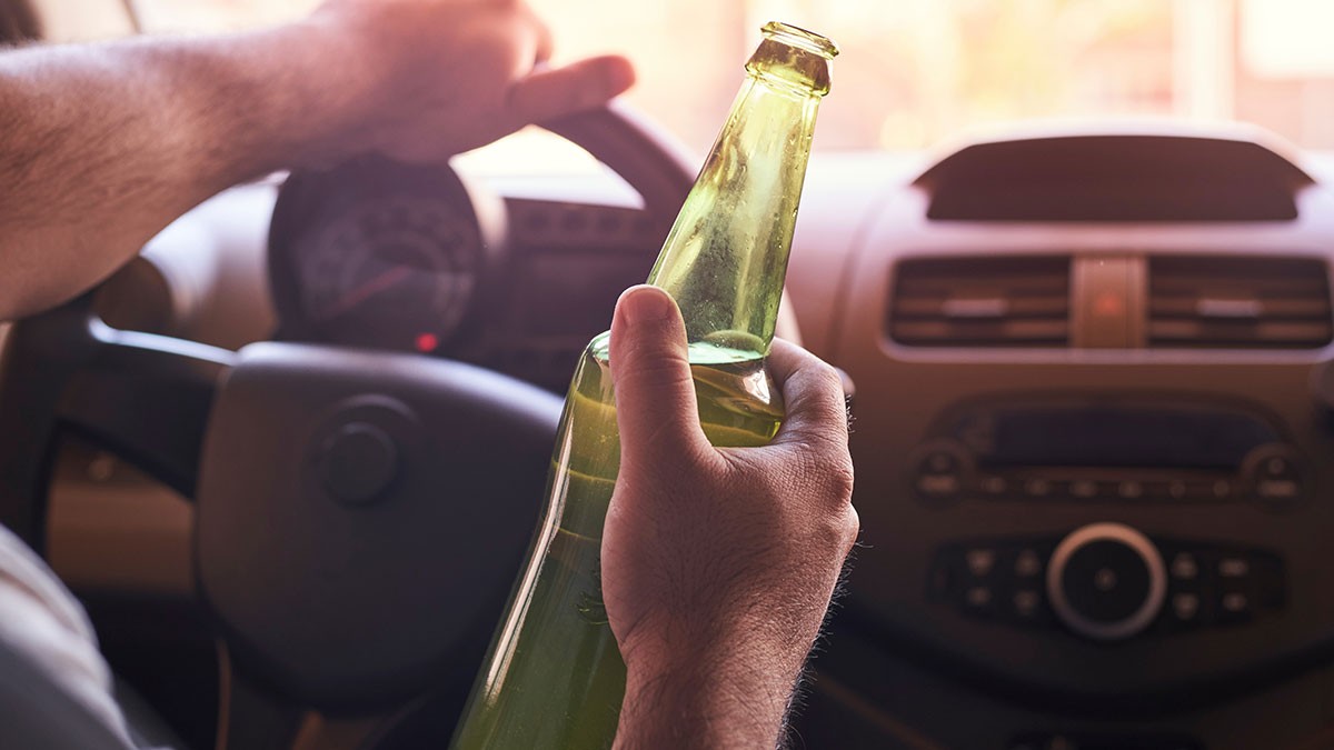 Person holding a beer bottle while driving, with one hand on the steering wheel inside a car.