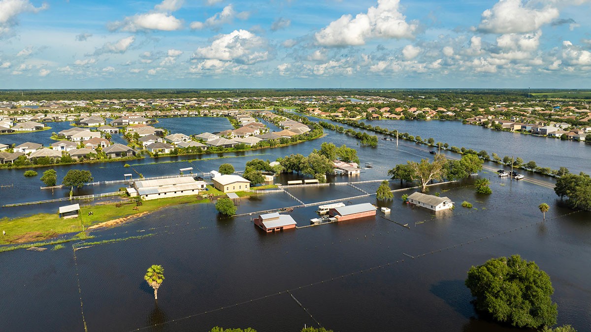 Aerial view of a flooded residential area with submerged houses, roads, and trees under a partly cloudy sky.