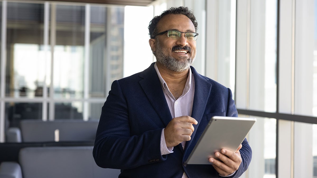 Smiling man in a navy blazer holding a tablet and stylus beside sunlit office windows.