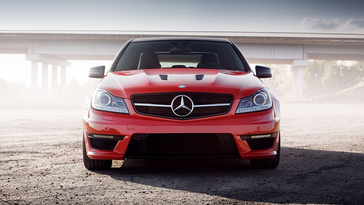 Front view of a red Mercedes-Benz sports car parked on a gravel surface, with a bridge and cloudy sky in the background.