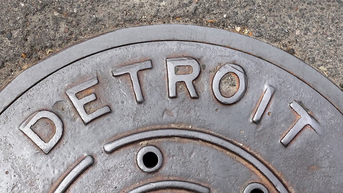 Close-up of a metal manhole cover with the word "DETROIT" embossed on it, set against a textured concrete surface.