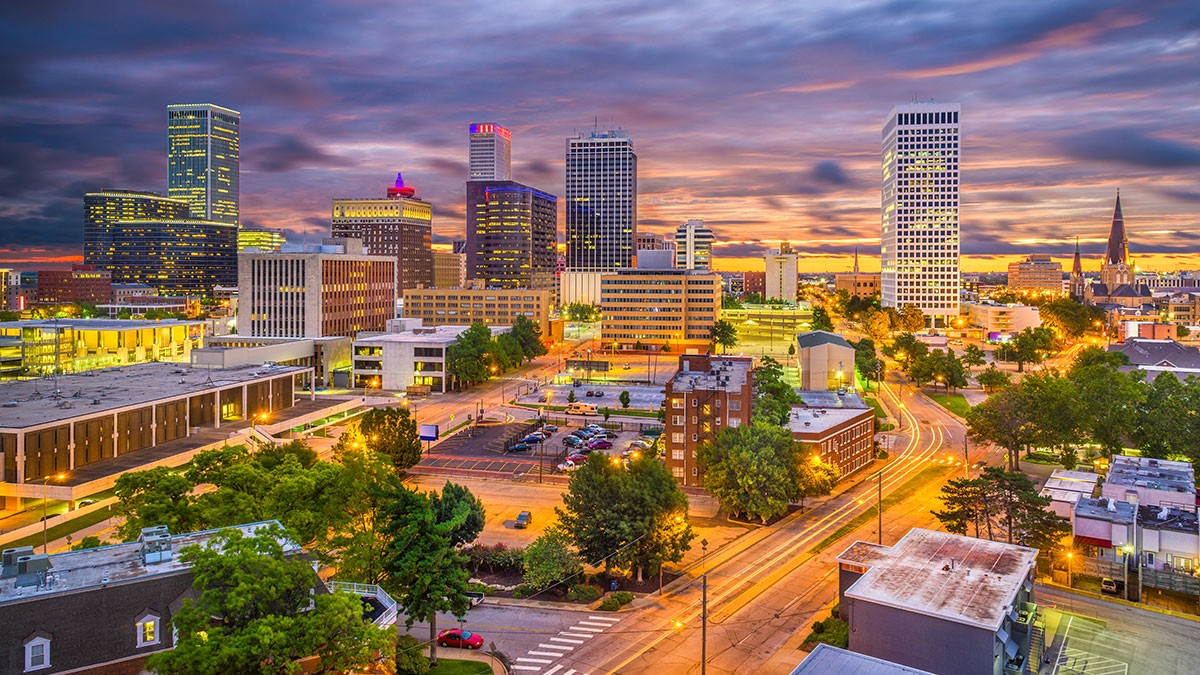 City skyline at dusk with illuminated buildings, vibrant sky, and roads lined with trees leading into the downtown area.
