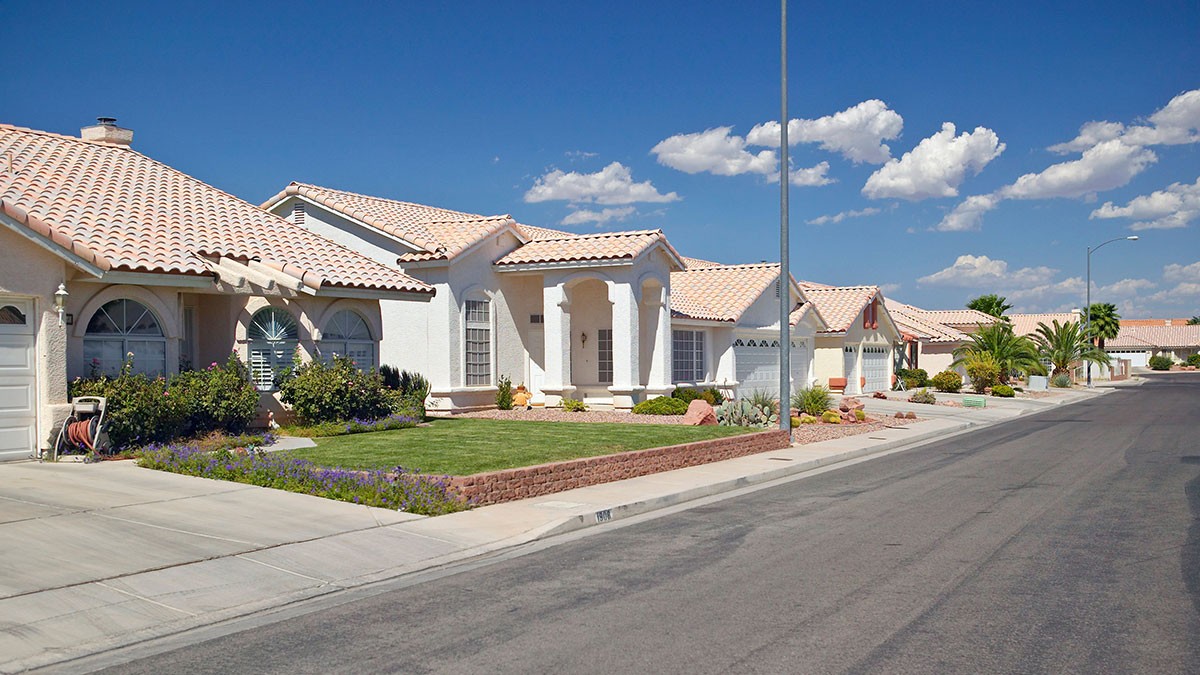 Suburban street with rows of similar white houses, red-tiled roofs, manicured lawns, under a clear blue sky with scattered clouds.