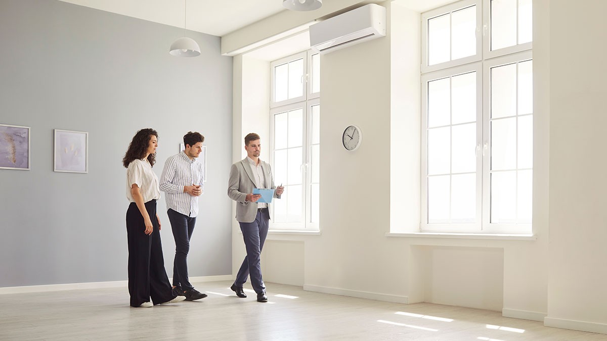 Three people in a bright, empty room with large windows, a wall clock, and an air conditioner. One person holds a clipboard.