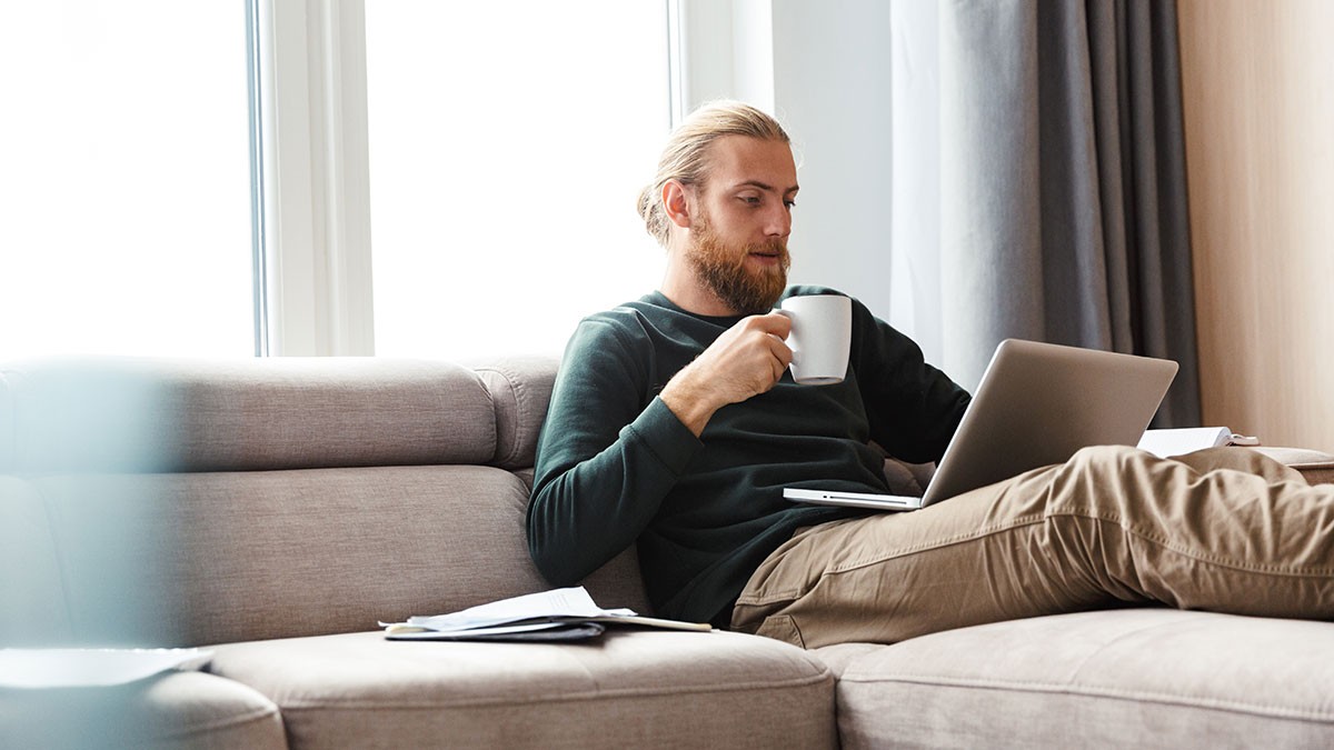 A man with a beard relaxes on a sofa, drinking from a mug and using a laptop, with papers scattered beside him.