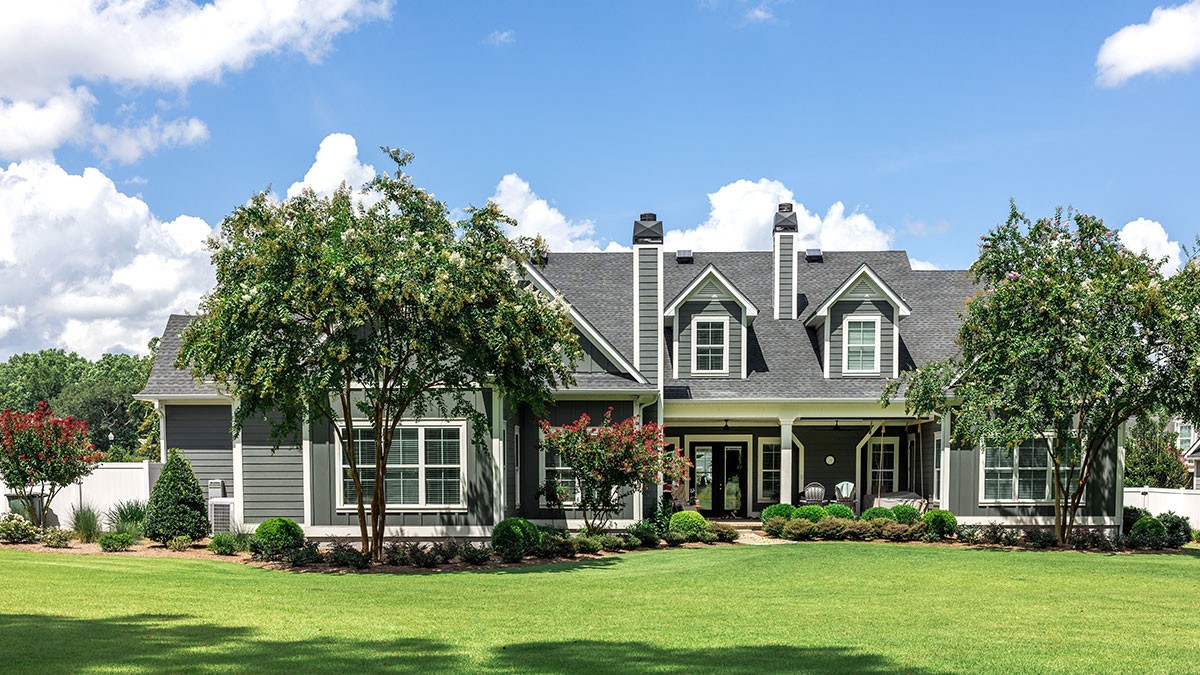 A large, modern house with gray siding, white trim, and a manicured lawn, surrounded by trees, under a partly cloudy blue sky.