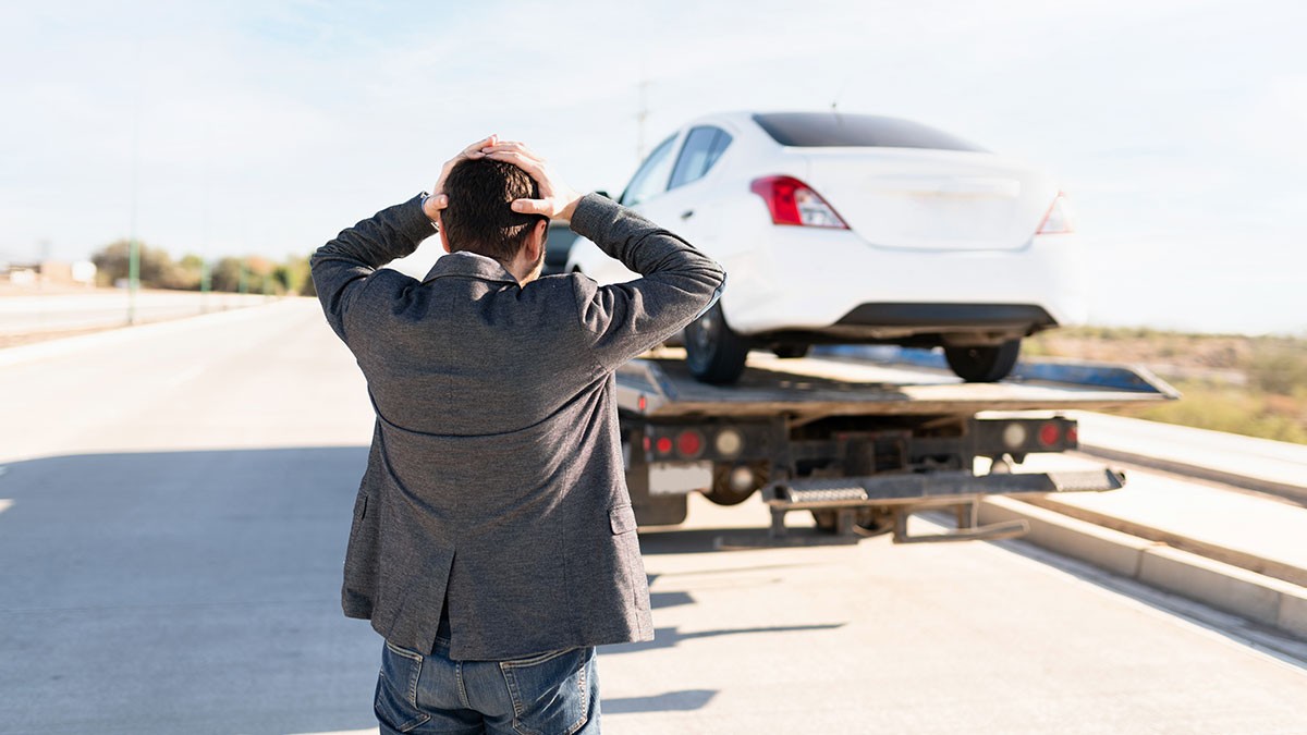 Man holding head in frustration as a white car is towed on a flatbed truck on an empty road.