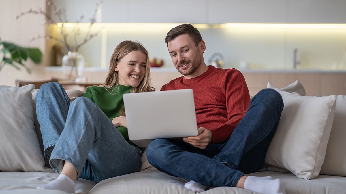 Couple sitting on a couch, smiling while looking at a laptop in a cozy living room setting with soft lighting.