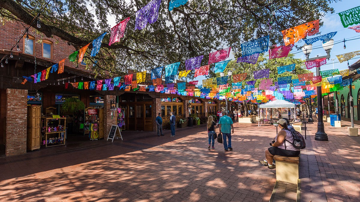 Festive outdoor market street with colorful papel picado banners hanging overhead, sunlight filtering through trees, and people walking or sitting.