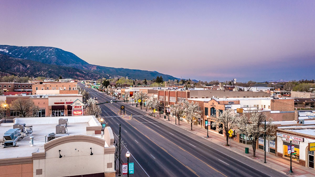 Aerial view of a small town at dusk, with a wide street lined with buildings and blossoming trees, and a mountain in the background.