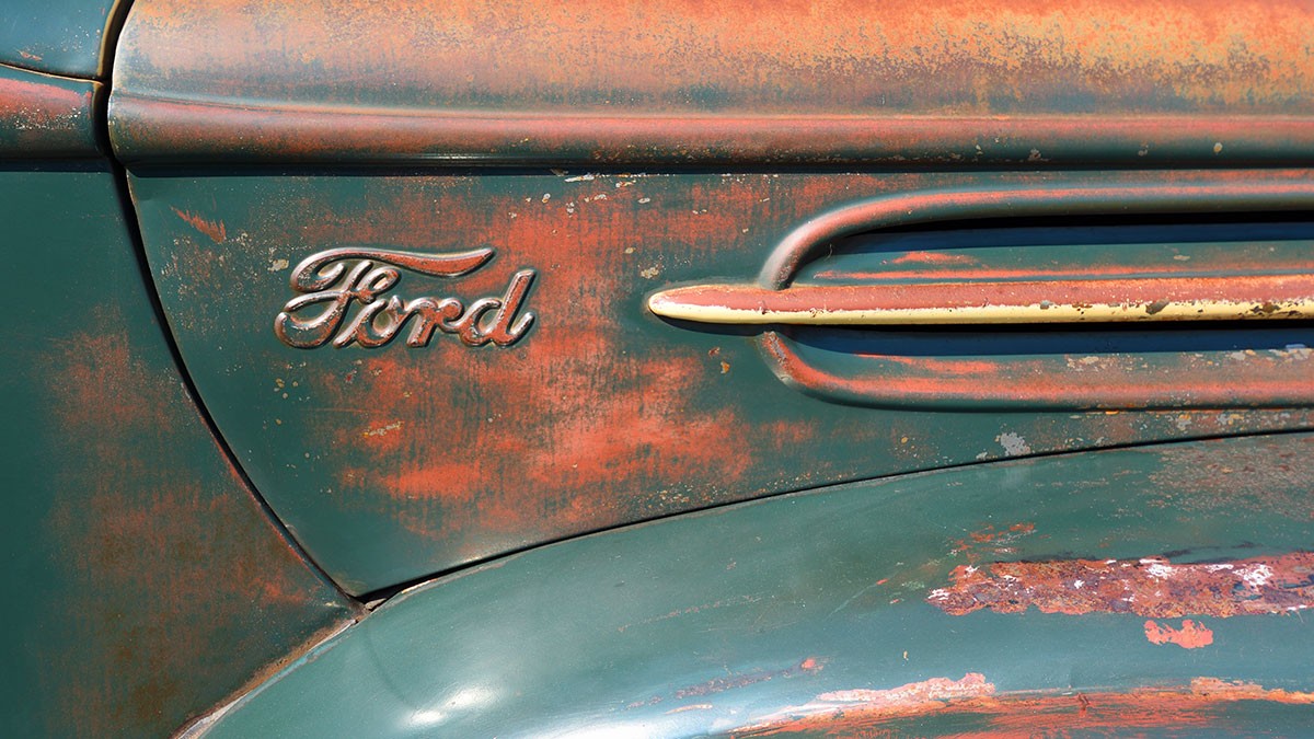 Close-up of a rusty vintage Ford car with visible logo and weathered paint, showcasing its aged and textured surface.