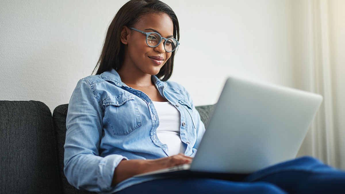 A woman wearing glasses and a denim shirt is sitting on a couch, smiling while using a laptop in a relaxed setting.