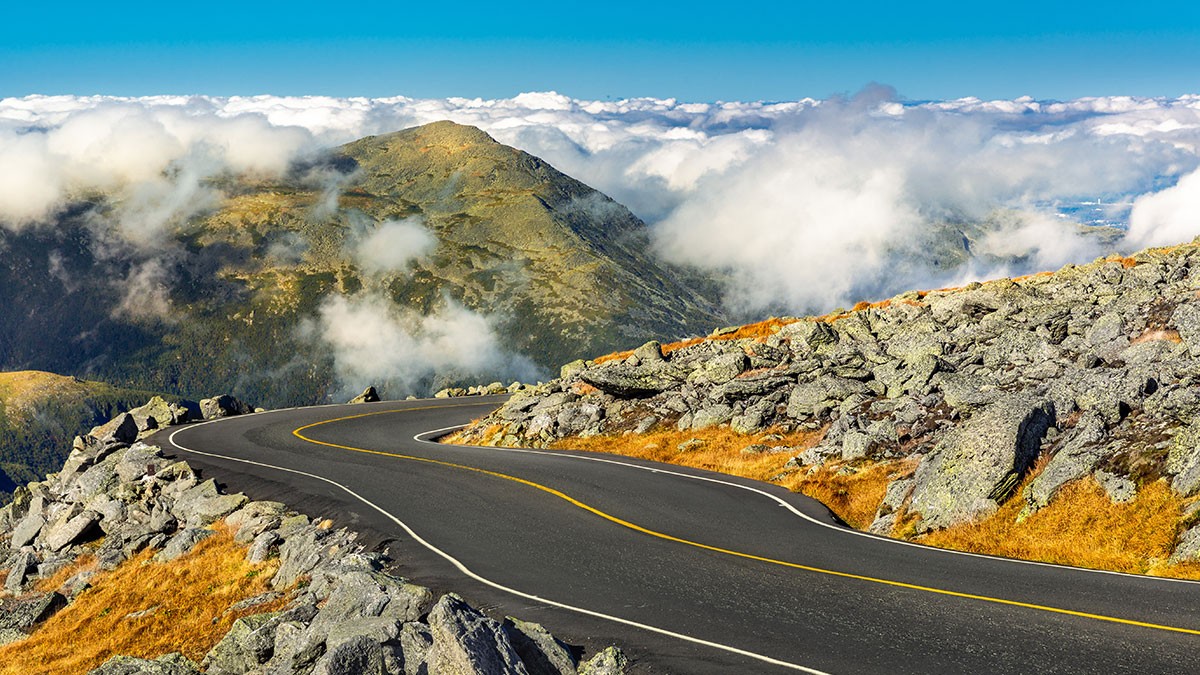 Winding mountain road with rocky edges, surrounded by clouds and a clear blue sky, leading towards distant peaks.