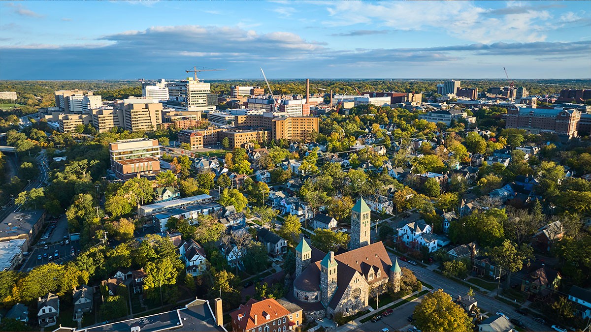Aerial view of a city with a mix of greenery, residential buildings, and taller city structures under a partly cloudy blue sky.