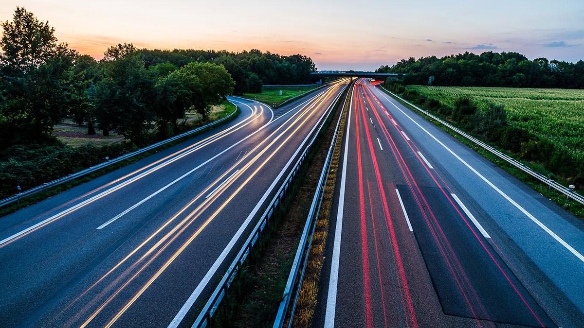 Aerial view of a highway at dusk with light trails from cars, surrounded by trees and fields under a colorful sky.
