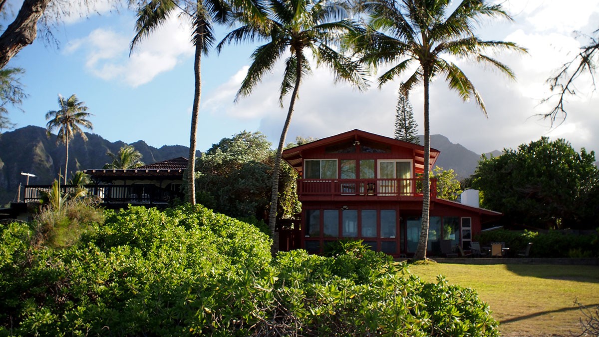Tropical house with tall palm trees, lush greenery, and mountains in the background under a blue sky with scattered clouds.