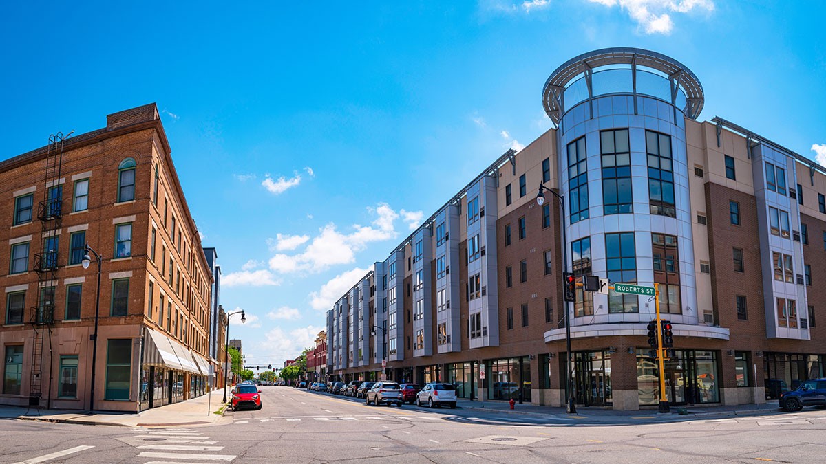 Sunlit intersection featuring a modern curved-glass mixed-use building across from an older brick commercial block, parked cars along the street.