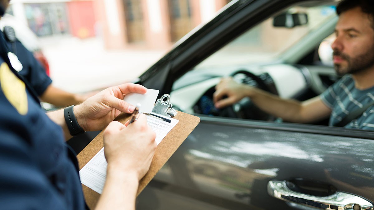 Police officer writing a ticket beside a car window, while the driver hands over an ID, parked on a city street.