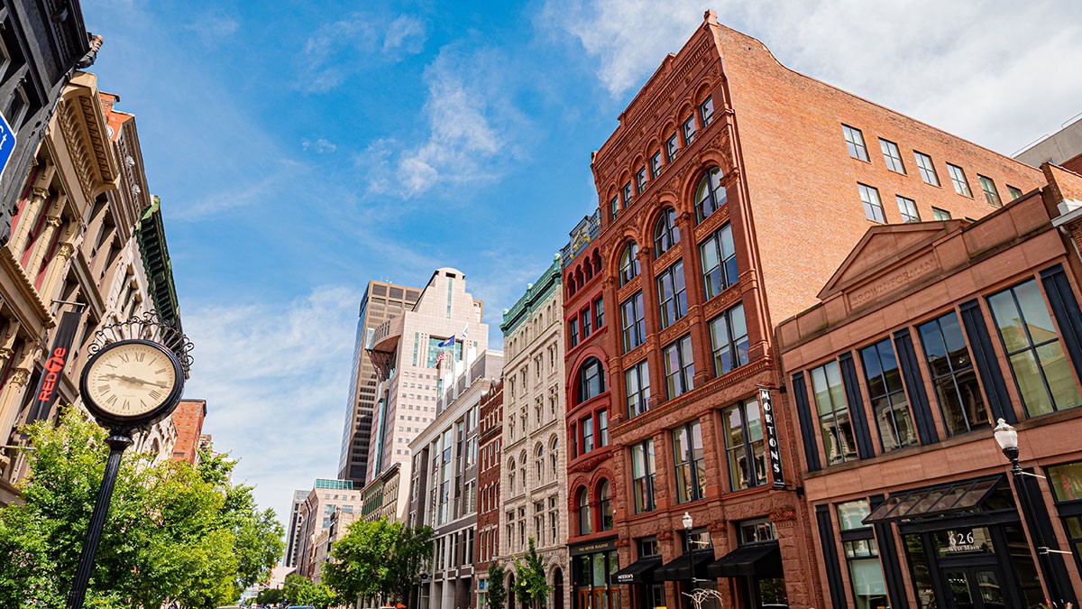 Street view with historic brick buildings, a vintage clock, and a clear blue sky. Lush green trees line the sidewalk.